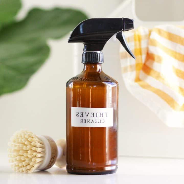 a glass amber spray bottle with a label that says thieves cleaner on it. There is a cleaning caddy, a striped rag and essential oil bottles in the background