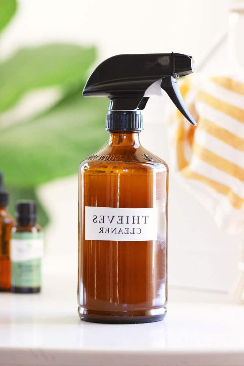a glass amber spray bottle with a label that says thieves cleaner on it. There is a cleaning caddy, a striped rag and essential oil bottles in the background