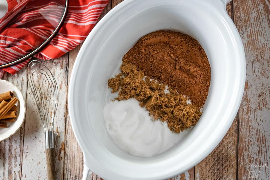 A white slow cooker with cocoa powder, brown sugar and granulated sugar next to a whisk.