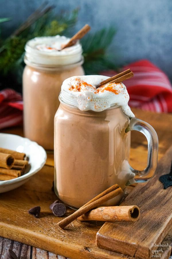 Clear glass mugs of Mexican Hot Cocoa topped with whipped cream and cinnamon sticks on a wooden tray.