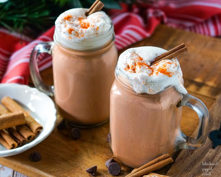 Two mugs of hot cocoa topped with whipped cream, cinnamon sticks, and cayenne pepper.