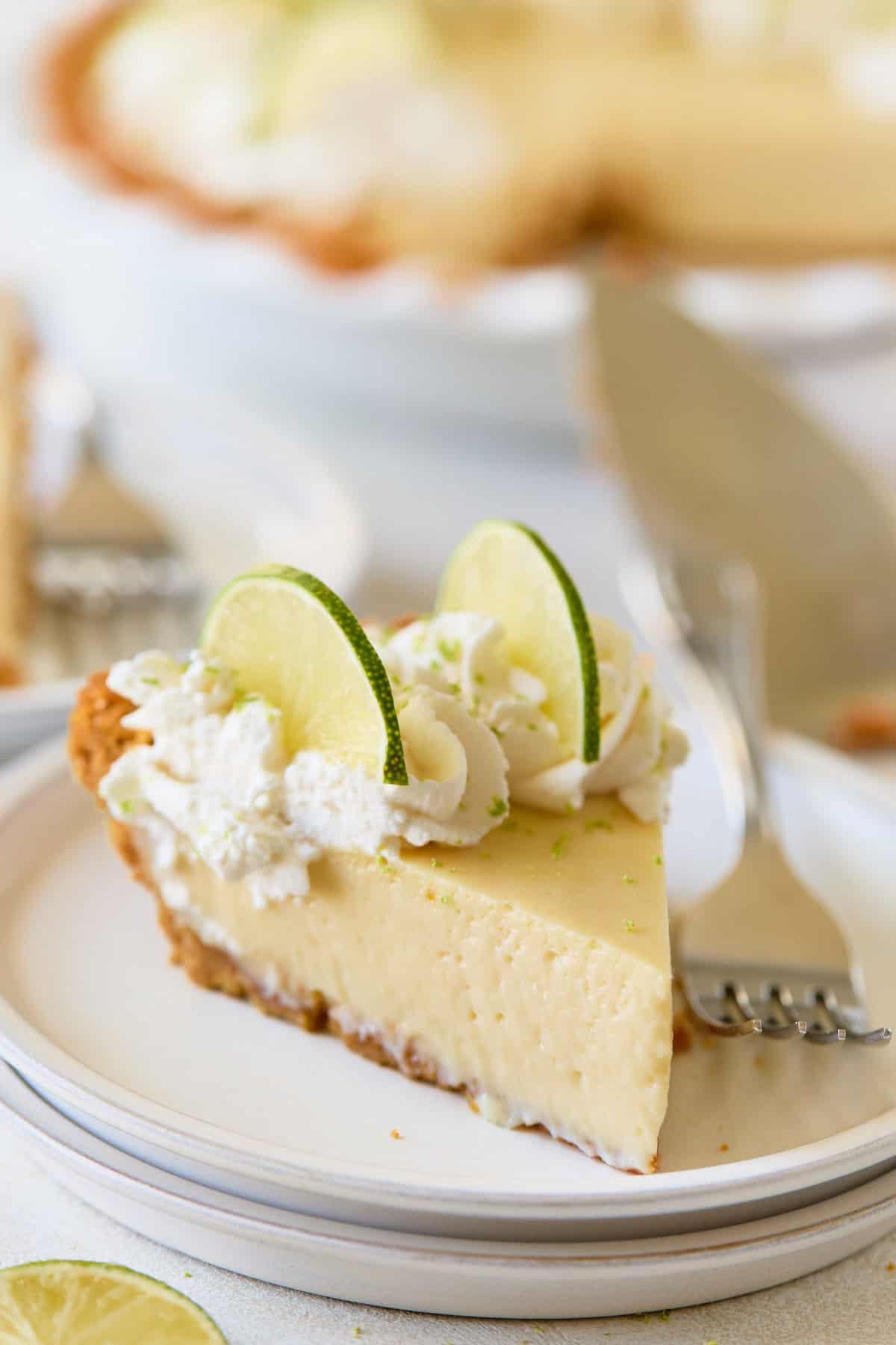 A close-up slice of creamy pie topped with whipped cream and lime slices on a white plate, with the whole pie in the background.