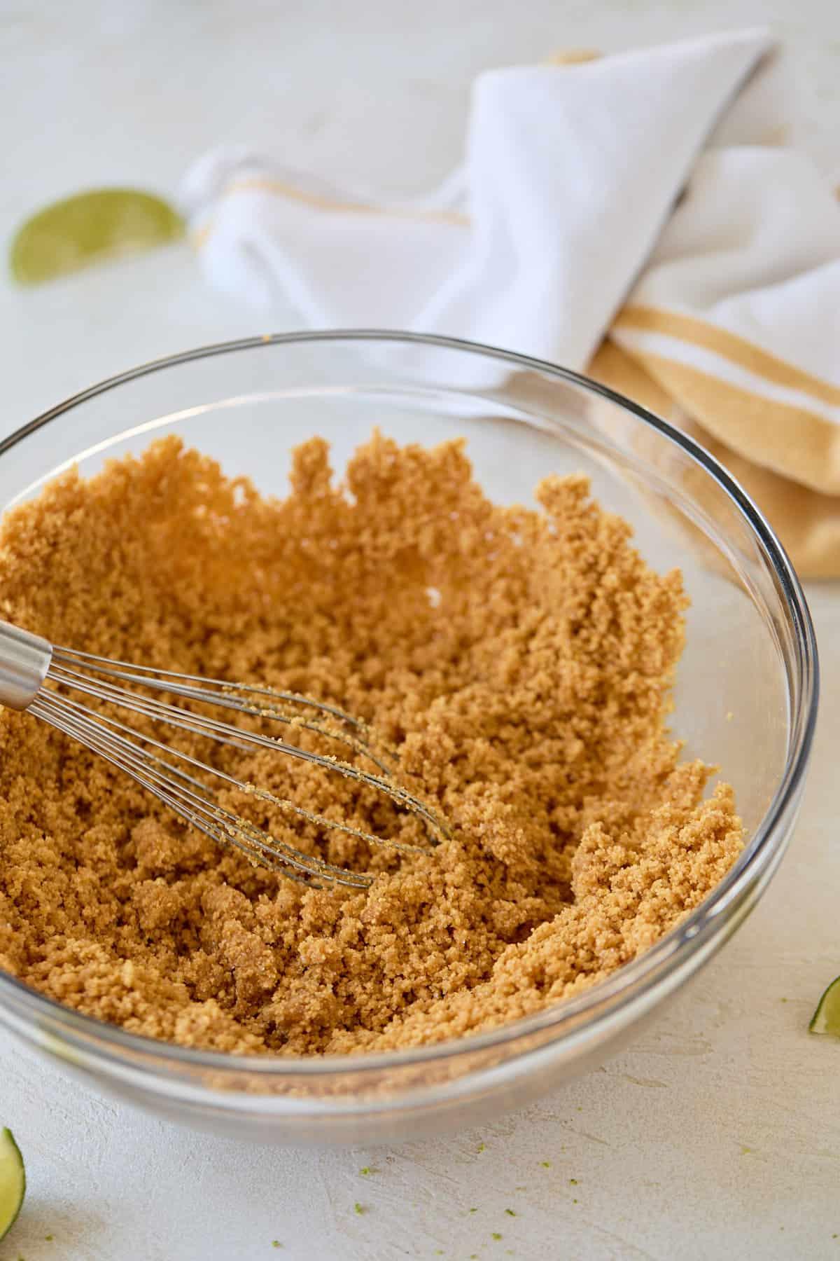 A clear glass mixing bowl filled with graham cracker crust mixture being whisked.