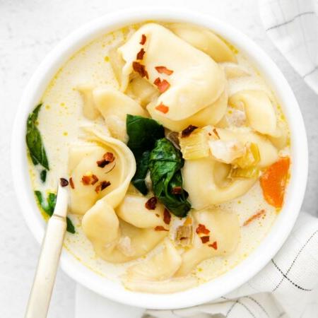 Photo of a bowl of Instant Pot Tortellini Soup that looks super rich and creamy with cheesy tortellini, fresh baby spinach, and carrots and garnished with a sprinkle of red pepper flakes